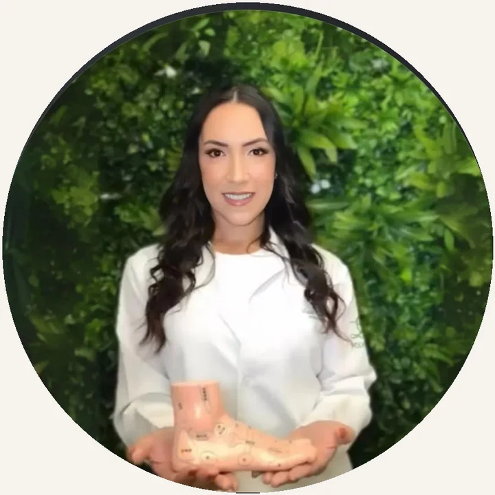 Laís Cardoso smiling in a white coat, holding a foot model, in front of a green plant wall